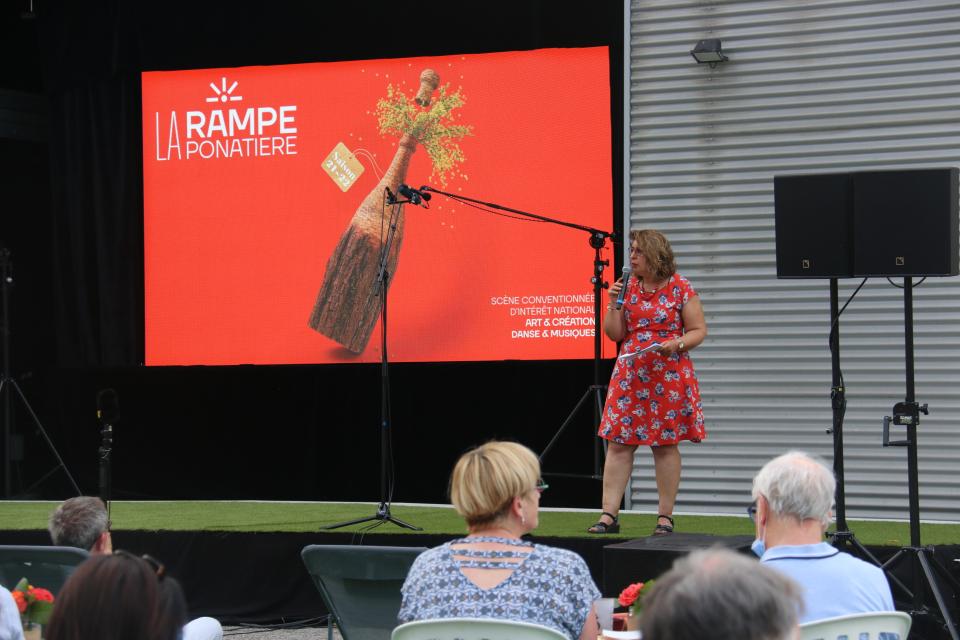 Photo de la présentation de saison la Rampe-La Ponatière pour la saison 2021-2022. Celle-ci se déroulait en extérieur. Nous voyons Joséfa Gallardo, directrice de La Rampe - La Ponatière, sur scène, un micro à la main, devant l'affiche de la saison.