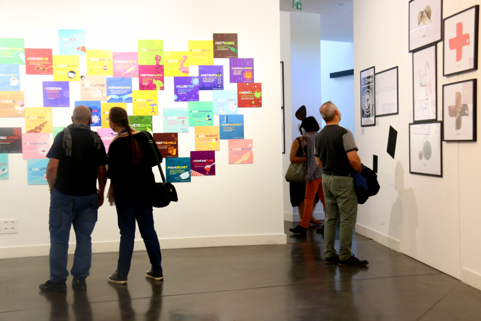 Des visiteurs devant Le lexique des confinés. Photo de l'exposition "L’art déconfiné des confinés" au centre du graphisme. Des visiteurs lisent le “Lexique des confinés”. Il s'agit de mini affiche très colorées et visuelles, expliquant certains " mots mêlés" comme "impuichiant".