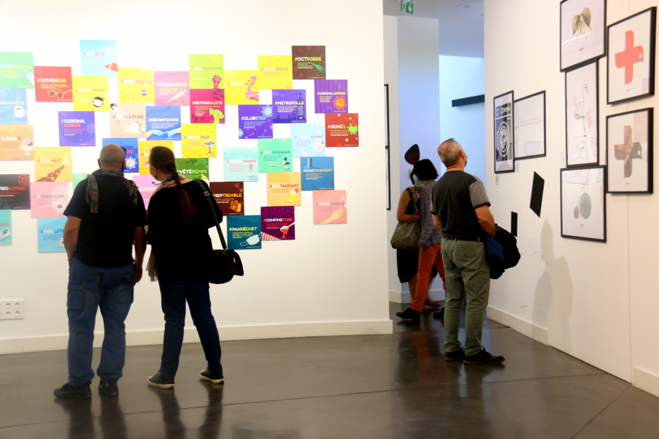 Photo de l'exposition "L’art déconfiné des confinés" au centre du graphisme. Des visiteurs lisent le “Lexique des confinés”. Il s'agit de mini affiche très colorées et visuelles, expliquant certains " mots mêlés" comme "impuichiant".