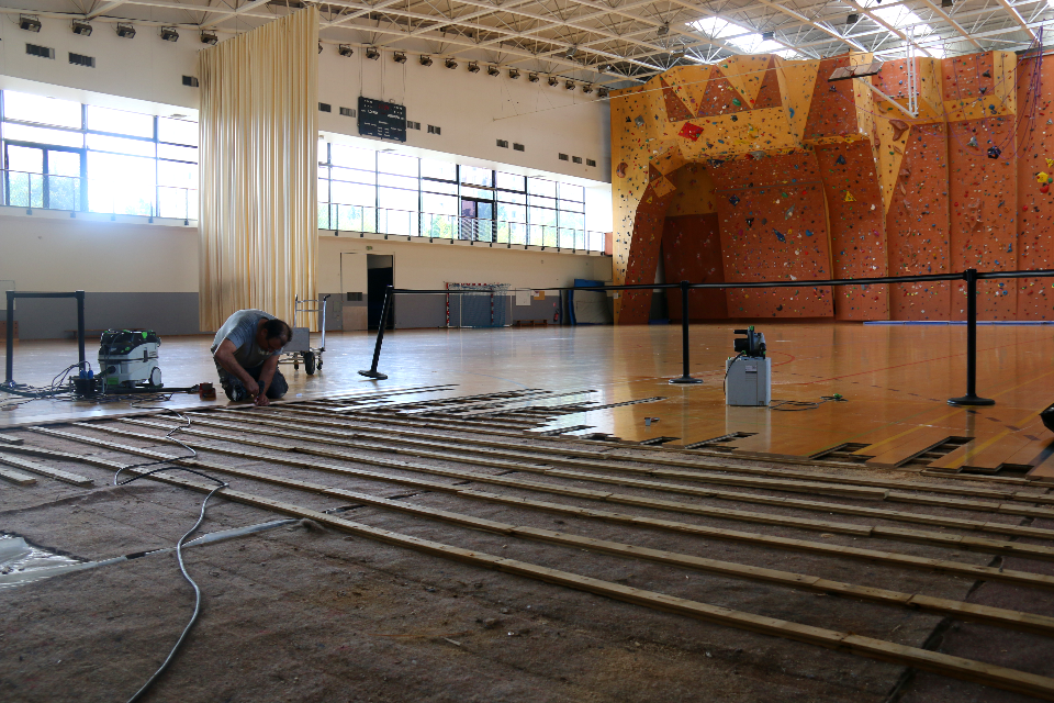 Photo des réparations du parquet du gymnase Lionel Terray. La photo est prise dans le gymnase, nous voyons le mur d'escalade en fond. Au premier plan nous voyons un technicien en train de changer les lames du sol.