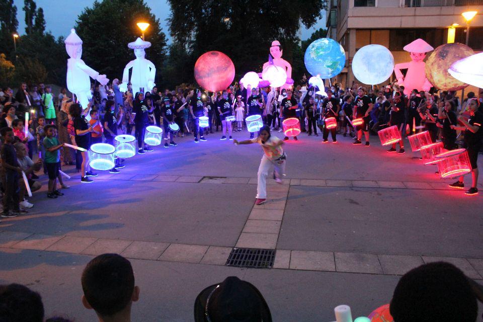 Photo prise à l'occasion de l'événement Carna’light.  Nous voyons des ballons géants lumineux en forme de boule, de baleine. des mascottes géantes en carton. Les participants sont habillés de vêtements blanc avec des marques fluorescentes dessus. Ils se reflètent dans la nuit.  Le public nombreux les entourent pour observer le spectacle. Au centre, un artiste fait une démonstration de hip hop